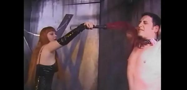 Two red-haired furies in latex dresses hang clothespins on the body of a young guy and spank him with a whip